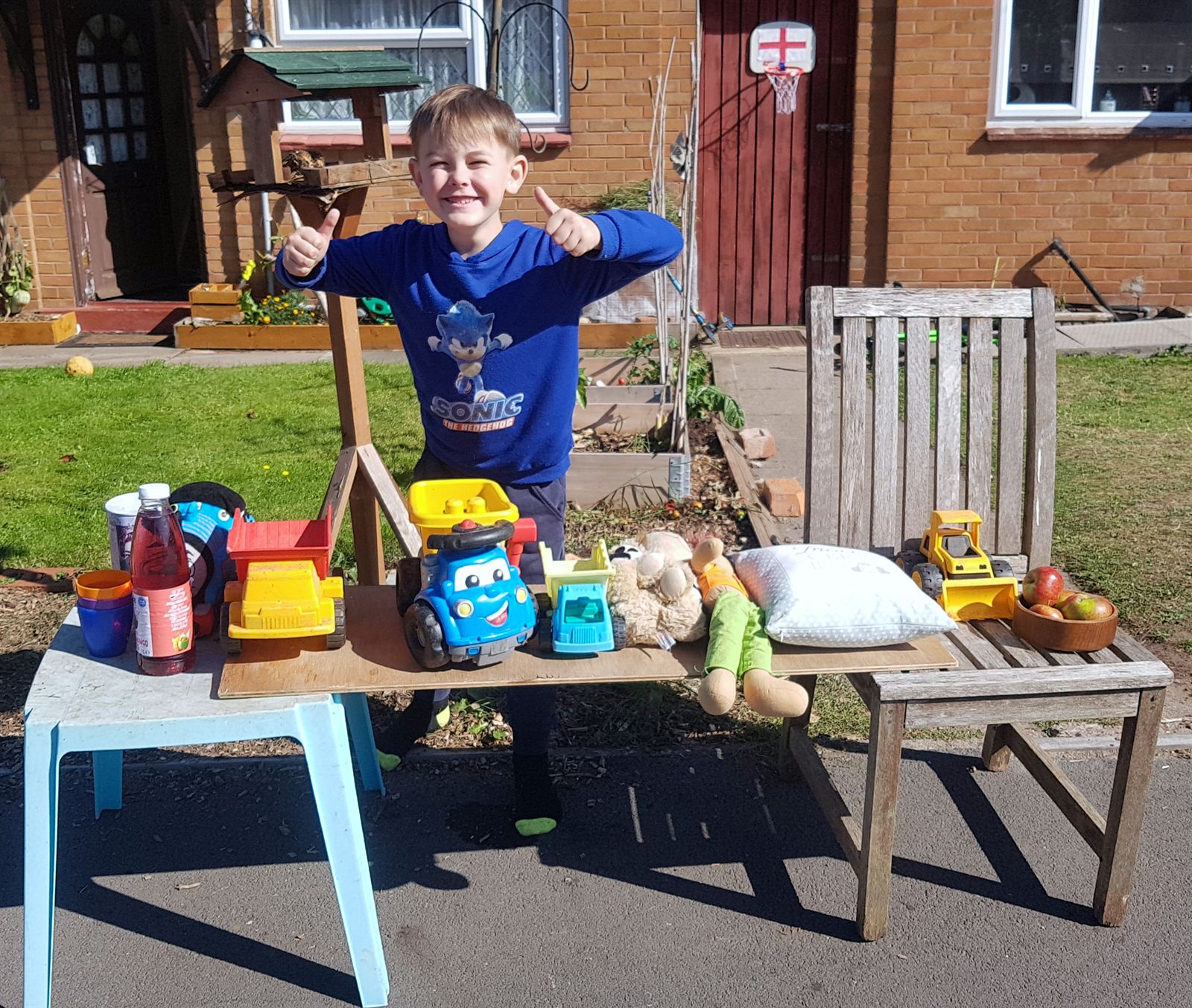 A boy giving two thumbs up behind a driveway table of toys and drinks for sale